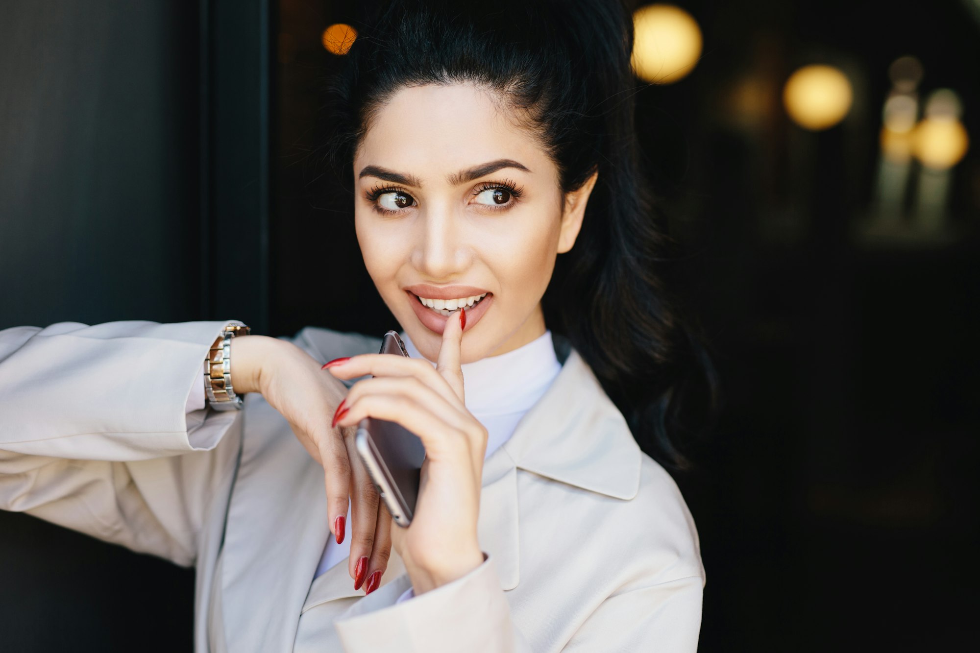 Adorable woman with appealing eyes, make-up, holding her finger on white teeth holding smartphone